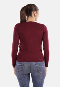Maglione a maniche lunghe bordeaux con design aderente, morbida texture e orlo a coste, abbinato a jeans in denim blu con tasche posteriori.