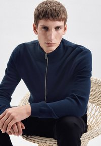 Marine blauwe ritstrui met een hoge kraag, geribbelde manchetten en een gladde textuur, gedragen door een model dat in een geweven stoel zit.