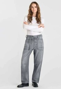 Jean gris à jambes larges et taille haute, avec de grandes poches avant et des coutures apparentes. Porté avec un haut blanc à manches longues et des chaussures noires.