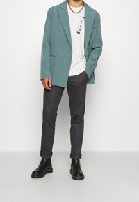 Tyrkysový blazer přes bílý tričko s potiskem, sladěný s tmavými kalhotami a černými kotníkovými botami. Hladká tkanina, uvolněný střih a minimální detaily.