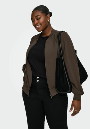 Smilende kvinne med mørk brun hettejakke med glidelås, svart topp, svarte bukser, bærer stor svart handlenett på skulderen, står foran hvit bakgrunn.