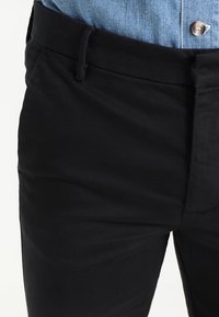 Černé bavlněné kalhoty s hladkou texturou, vybavené zipovým zapínáním, poutky na pásek a bočními kapsami, nošené s světle modrou džínovou košilí.