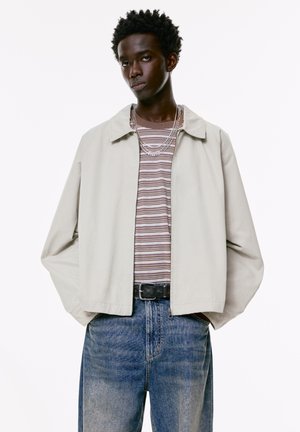 Jonge man in een lichtbeige jas, gestreept shirt, blauwe spijkerbroek, zwarte riem en meerdere zilveren kettingen, staand met zijn handen in zijn zakken.