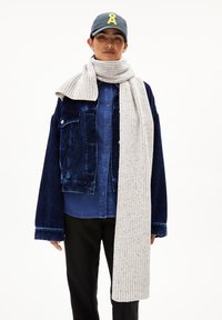 Marinefarbener Cordjacke über einem blauen Hemd mit Knöpfen, kombiniert mit einem strukturierten, hellgrauen Rippenschal. Schwarze Hosen und eine dunkle Kappe mit einem Logo.