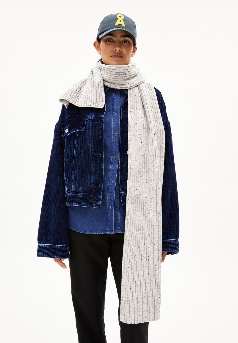 Marinefarbener Cordjacke über einem blauen Hemd mit Knöpfen, kombiniert mit einem strukturierten, hellgrauen Rippenschal. Schwarze Hosen und eine dunkle Kappe mit einem Logo.