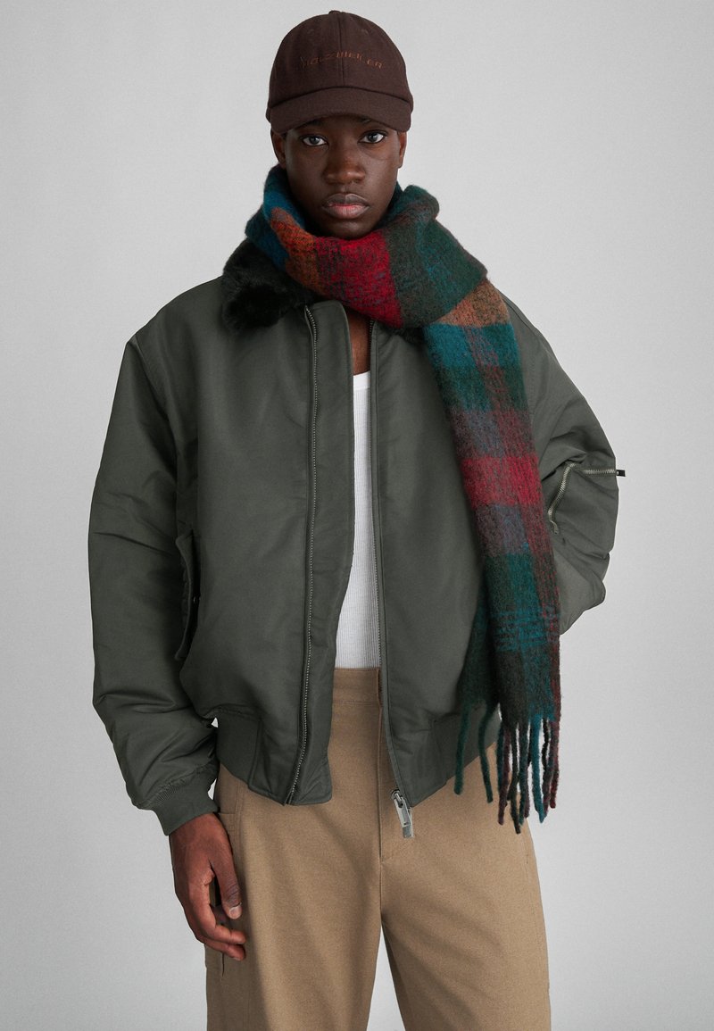 Oļveļzaļš bomberjaka ar ribotiem aprocēm, kopā ar daudzkrāsainu plecu šalli sarkanos, zilās un zaļās toņos, un bēša krāsas biksēm.