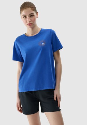 Młoda kobieta ubrana w niebieski t-shirt z krótkim rękawem z małym nadrukiem z przodu i czarne krótkie spodenki sportowe, stojąca na jednolitym tle.