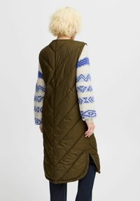 Langer olivgrüner Steppweste mit Zickzacksteppung, seitlichen Schlitzern und einem kontrastierenden, gemusterten Pullovern mit Ärmeln, die blaue geometrische Designs aufweisen.