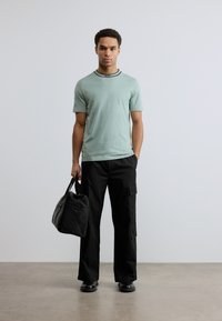 Ανοιχτό πράσινο βαμβακερό t-shirt με μαύρο ριμπ λαιμό, συνοδευόμενο από μαύρο cargo παντελόνι με πλαϊνές τσέπες και μαύρη τσάντα ώμου.