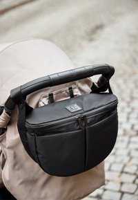 Musta vetoketjullinen lastenvaunujen järjestäjä kiinnitettynä beigen väriseen lastenvaunukahvaan mukulakivikadun taustalla.