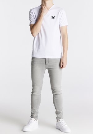 Hvid bomulds t-shirt med v-hals og sort logo, parret med lysegrå slim-fit jeans og hvide sneakers. Simpelt, afslappet outfit-design.