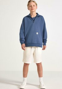 Blå fleece zip-up hoodie med framficka, i kombination med ljusa shorts och vita sneakers. Avslappnad, luftig design.
