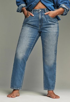 Person iført højtkalvende, falmede blå jeans og en denimjakke med hænderne i lommerne, stående barfodet mod en ensfarvet baggrund.