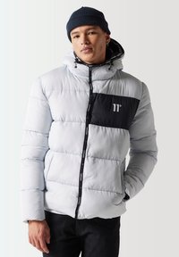 Chaqueta de plumas gris claro con un bolsillo en el pecho negro, que cuenta con capucha, cierre frontal con cremallera y tejido texturizado. Incluye detalles de cordón ajustable.