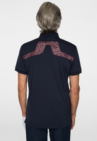 Granatowy polo z teksturowanym bordowym wzorem na ramionach, krótkimi rękawami i prostym dołem. Materiał wydaje się być oddychający i miękki.