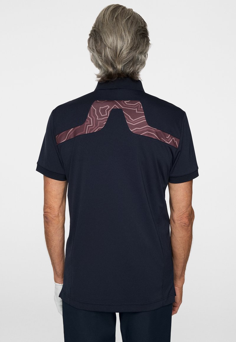 Granatowy polo z teksturowanym bordowym wzorem na ramionach, krótkimi rękawami i prostym dołem. Materiał wydaje się być oddychający i miękki.