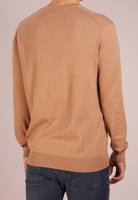 Person i en kamelfärgad långärmad tröja och mörkblå jeans, sedd bakifrån mot en enkel ljus bakgrund.