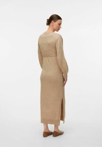 Abito beige lavorato a maglia con maniche lunghe, vita aderente e spacco laterale. Texture morbida, lunghezza fino al polpaccio, rifinito con orlo a costine. Scarpe marroni senza lacci.