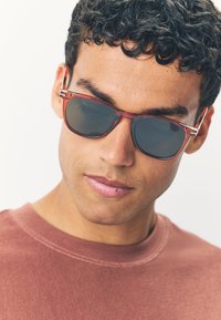 Jonge man met krullend haar, donkere zonnebril en een gedempt rood shirt met ronde hals, die iets naar beneden kijkt met een neutrale uitdrukking.