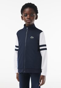 Granatowa kurtka z zamkiem błyskawicznym z białymi długimi rękawami, dwoma granatowymi paskami na rękawach, logo Lacoste na klatce piersiowej, wykonana z miękkiego, elastycznego materiału.