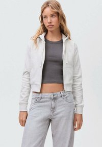 Felpa grigia con cappuccio a costine e tasche frontali, indossata sopra un top scuro aderente, abbinata a jeans leggeri a vita alta grigi.