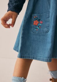 Robe en denim avec une jupe évasée, dotée d'une poche brodée avec un motif floral en orange, rose et vert. Tissu texturé bleu.