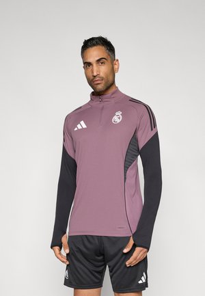 Paarse sporttop met een halve rits, zwarte mouwen en witte Adidas-strepen. Voorzien van het Real Madrid-logo en gestructureerde zijkanten.