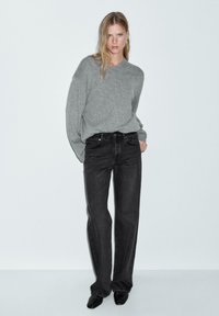 Maglione grigio a maglia con maniche leggermente oversize, abbinato a jeans neri a vita alta. Il modello si trova in piedi con le mani nelle tasche, indossando scarpe nere a punta.