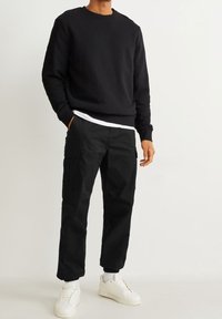 Homme portant un sweat à col rond noir, un pantalon cargo noir, des baskets blanches et des chaussettes blanches, debout avec une main dans la poche.
