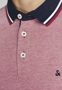 Rød tekstureret polo skjorte med navy krave, der har to hvide striber, tre knaplukninger og et lille mørkt logo på brystet.