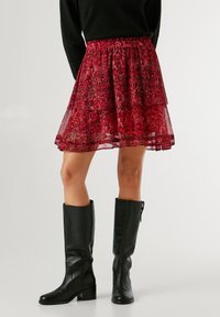 Punainen kuvioitu hame, jossa on kerroksellinen muotoilu, joustava vyötärö ja puoliläpinäkyvä rakenne. Yhdistetty polven ylle ulottuviin mustiin nahkasaappaisiin.