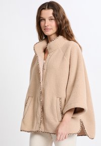 Beżowy polarowy poncho z wykończeniem w panterkę, bez kołnierza, z bocznymi kieszeniami oraz luźnym krojem. Miękka tekstura i przytulny wygląd.