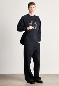 Tumši zils džemperis arapaļas formas logotipa detaļu, valkāts kopā ar vaļējām tumšām džinsa biksēm. Modelis tur melnu plecu somu, stāvot uz neitrālas krāsas fona.