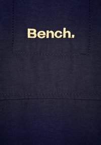 Námořnicky modrá látka s vroubkovanou texturou obsahuje našitou nášivku s výšivkou "Bench." v světle béžové barvě. Jednoduchý design bez dalších detailů.