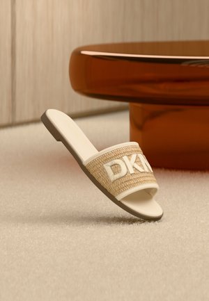 Beige Sandale mit Riemen aus geflochtenem Material und weißem DKNY-Logo, auf einem hellen Teppich neben einem runden, bernsteinfarbenen Tisch platziert.