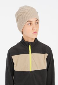 Beige gerippte Strickmütze, getragen mit einer schwarzen Fleecejacke, die ein beiges horizontales Band und einen leuchtend gelben Reißverschluss aufweist, was den texturalen Kontrast betont.