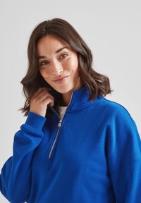 Blue zip-up sweatshirt made of soft fabric, featuring a high collar and ribbed cuffs, with a visible zipper detail at the neck.