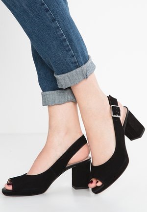 Feet in rolled-up blue jeans wearing black suede open-toe slingback block-heeled shoes with silver buckle.