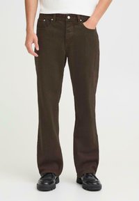 Mørkebrune denimjeans med rett passform, fem lommer og knappelukking. Teksturert stoff synlig; brukt med svarte sko.
