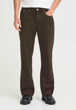 Mørkebrune denimjeans med rett passform, fem lommer og knappelukking. Teksturert stoff synlig; brukt med svarte sko.