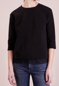 Svart sweatshirt med trekvartsärmar, ribbat mönster och spetsdetaljer vid fållen, parat med blå denimjeans.