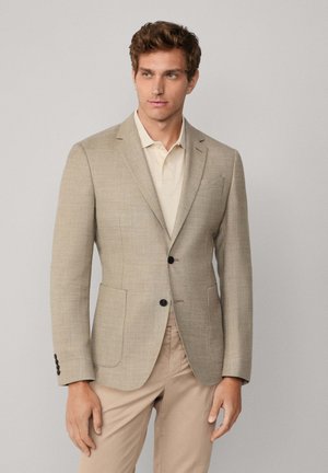 Beige Blazer mit strukturierter Webart, mit Reverskragen, zwei vorderen Taschen und dunklen Knöpfen, kombiniert mit einem hellen Poloshirt.