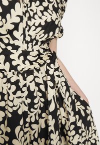 Robe avec motif abstrait de feuilles noir et beige, taille cintrée avec détail nœud, et jupe plissée, montrée de la taille jusqu'à mi-cuisse.