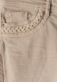 Beige denim kangas, joka sisältää punottuja yksityiskohtia taskureunassa, sileä pinta ja klassinen viisi-taskuinen muotoilu.
