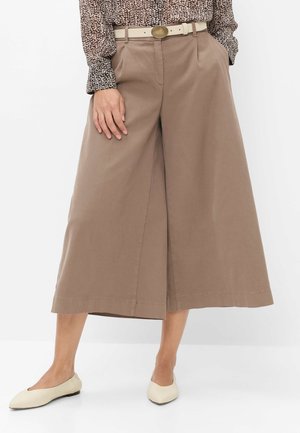 Vrouw draagt beige brede cropped broek, een blouse met lange mouwen en patroon, een witte riem met gouden gesp en crèmekleurige platte schoenen.