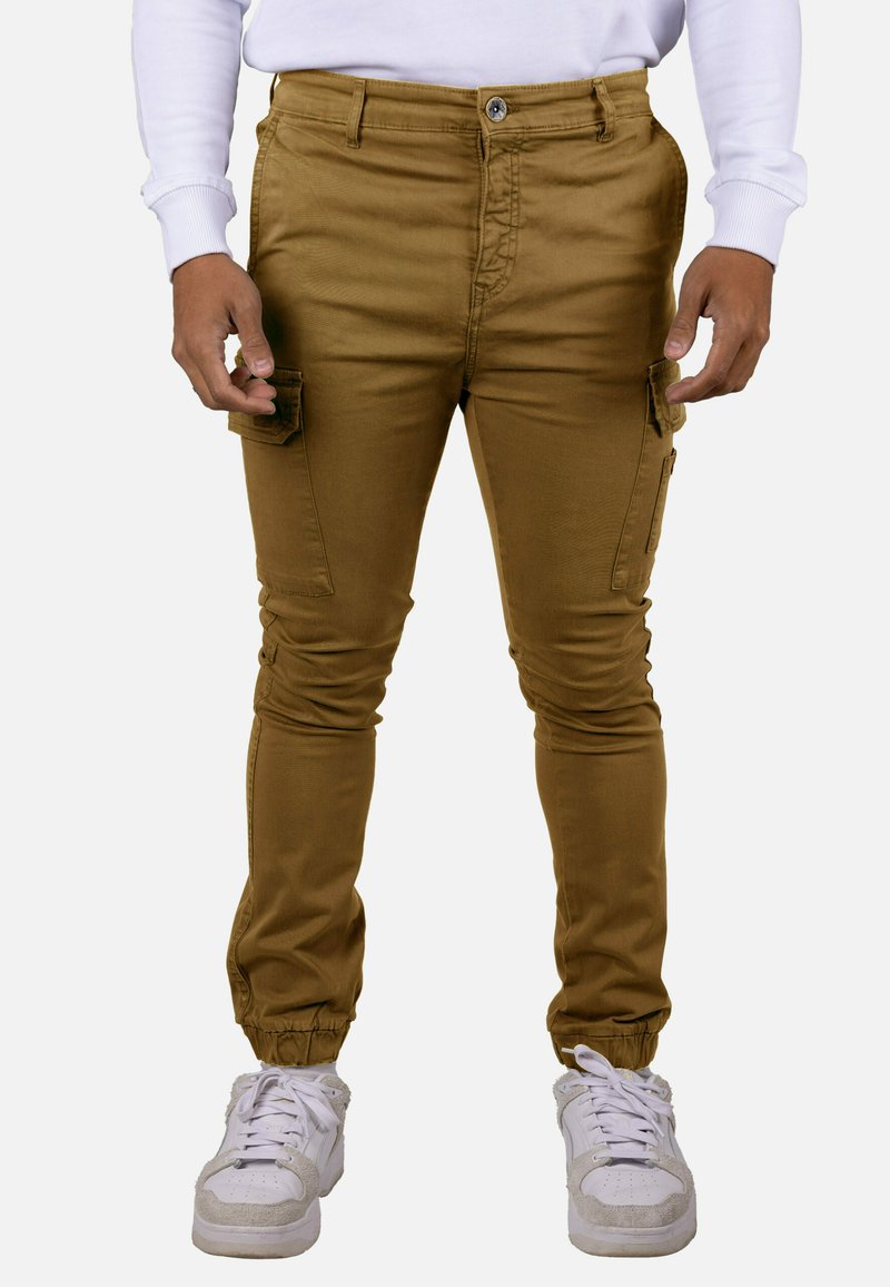 Pantalones cargo marrones de corte ajustado, tobillos estrechos, bolsillos laterales y cierre de botón. Combinados con zapatillas blancas y una camisa de manga larga.