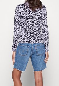 Fioletowa bluzka z długim rękawem w czarny wzór panterki, połączona z niebieskimi szortami dżinsowymi o postrzępionych brzegach i kieszeniach.
