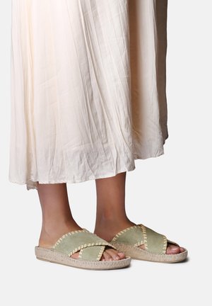 Pėdos dėvi smėlinės spalvos lengvus sandalus su kryžminiais dirželiais ir siuvimo detalėmis, derinamus su šviesia, klostuota midi ilgio suknele.