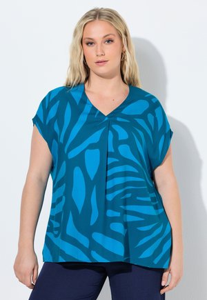 Frau mit langem blondem Haar trägt ein lockeres blaues Oberteil mit abstraktem, dunklerem blauem Muster und dunkler Hose, sie steht vor hellem Hintergrund.