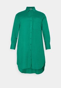 Grön långärmad skjorta, knapplång, med krage och en enkel bröstficka. Slät textur, rundad fåll och något oversized passform.